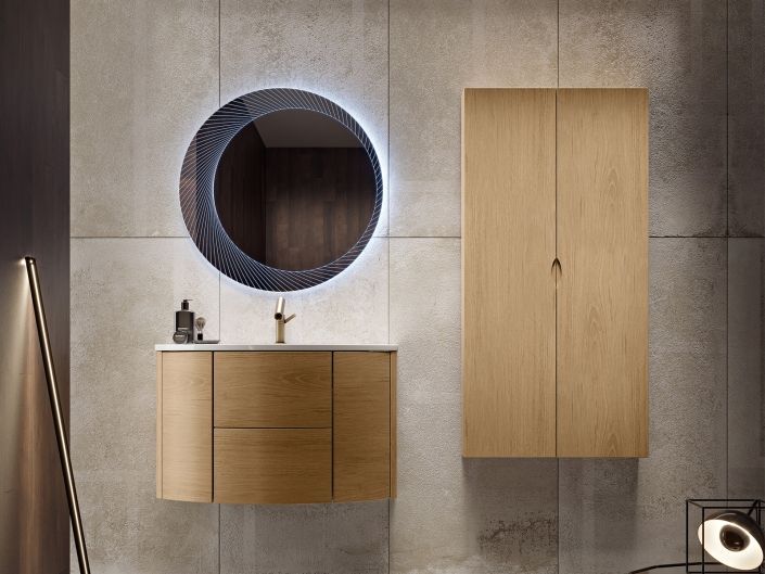 Bagno moderno con mobile lavabo in legno, specchio rotondo e pensile a muro, su pareti rivestite in piastrelle di cemento.