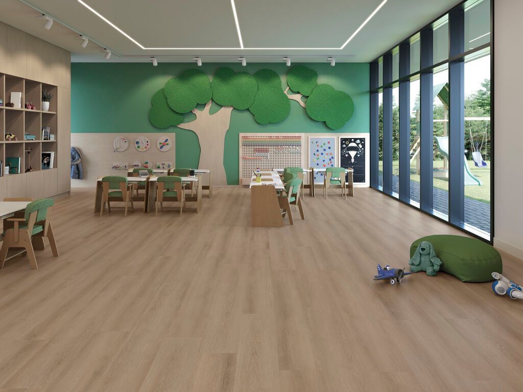 Aula luminosa con tavoli in legno, un murale raffigurante un albero e grandi finestre, con giocattoli sparsi sul pavimento.