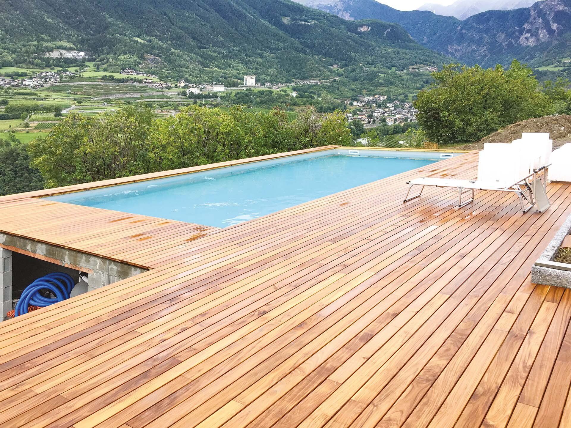 Terrazza in legno con piscina turchese affacciata su una verde vallata di montagna e lettini bianchi.