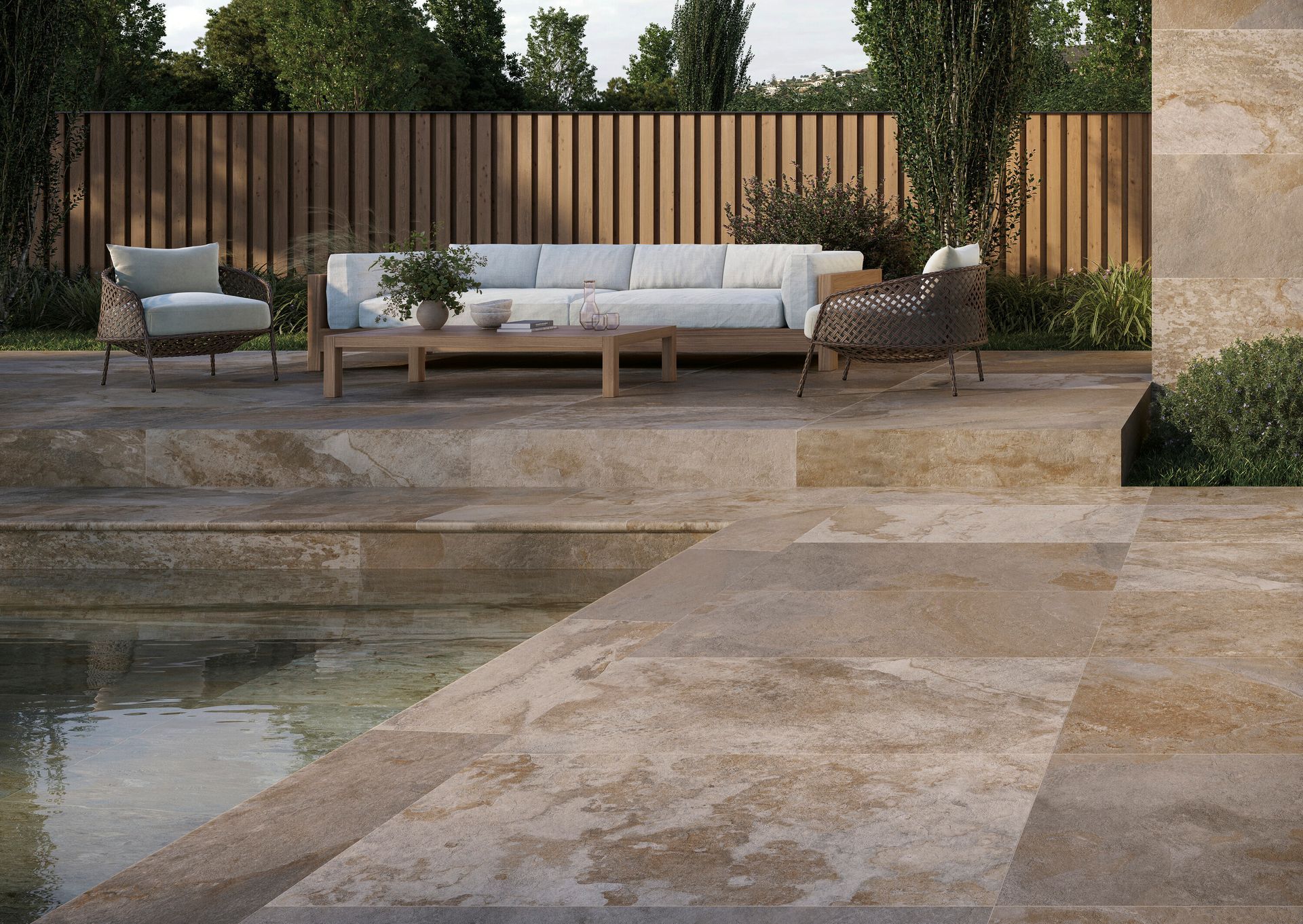 Patio moderno con pavimentazione in pietra, divano basso bianco, sedie e una piscina accanto a una recinzione in legno e vegetazione.