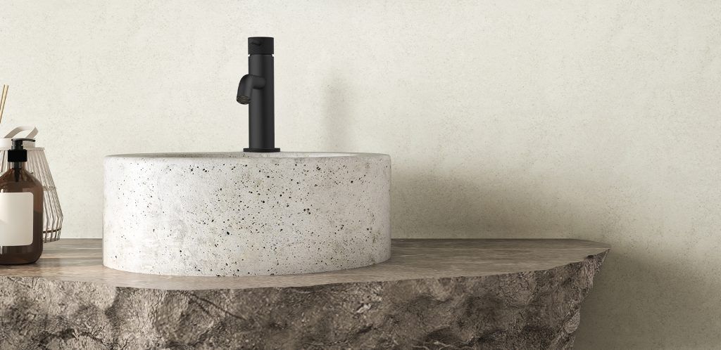 Lavabo in pietra con rubinetto nero su un piano in legno grezzo, addossato a una parete beige.