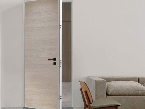 Porta di legno parzialmente aperta in una stanza minimalista con divano e poltrona visibili accanto.