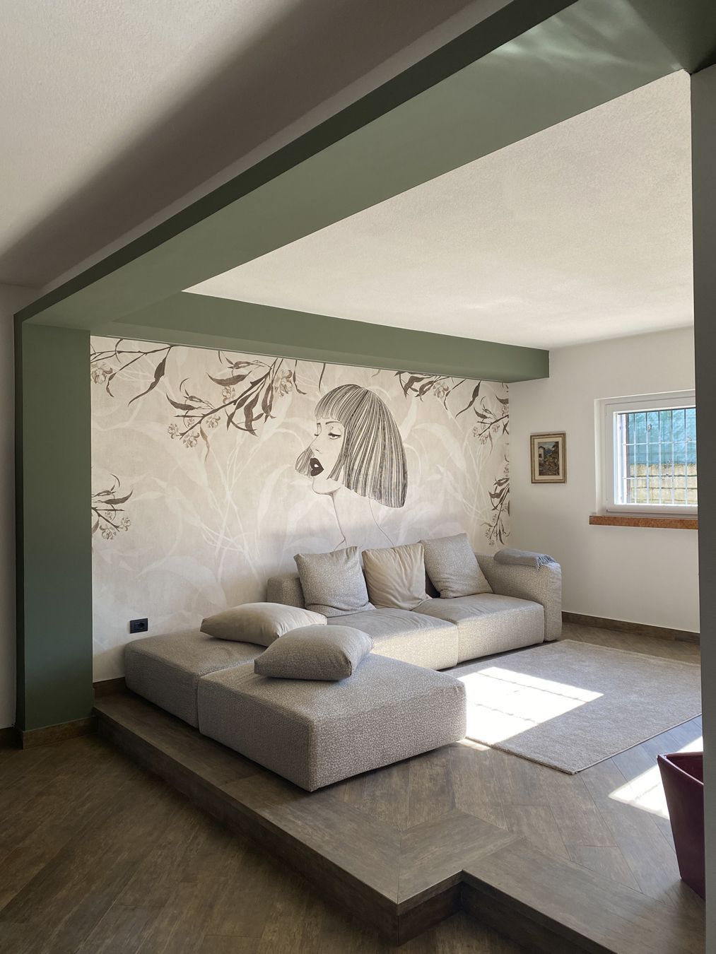 Soggiorno minimalista con divano angolare grigio chiaro, parete decorata con un murale e luce naturale proveniente da una finestra.
