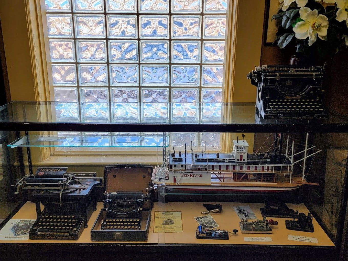 Three old typewriters are on display in a glass case in front of a window.