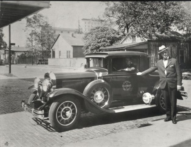 A black and white photo of a man standing next to an old car