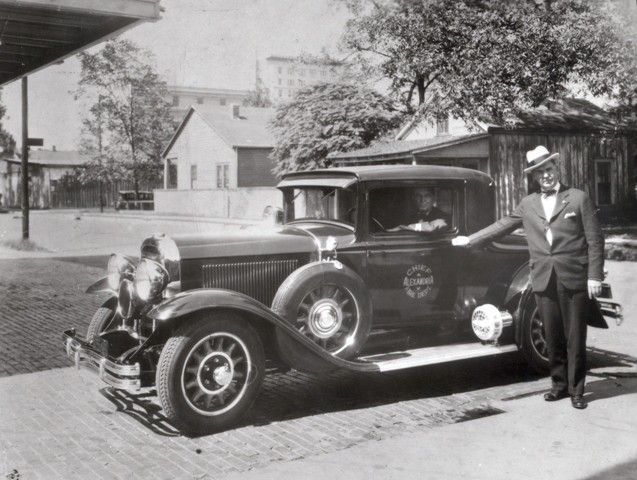 A black and white photo of a man standing next to an old car