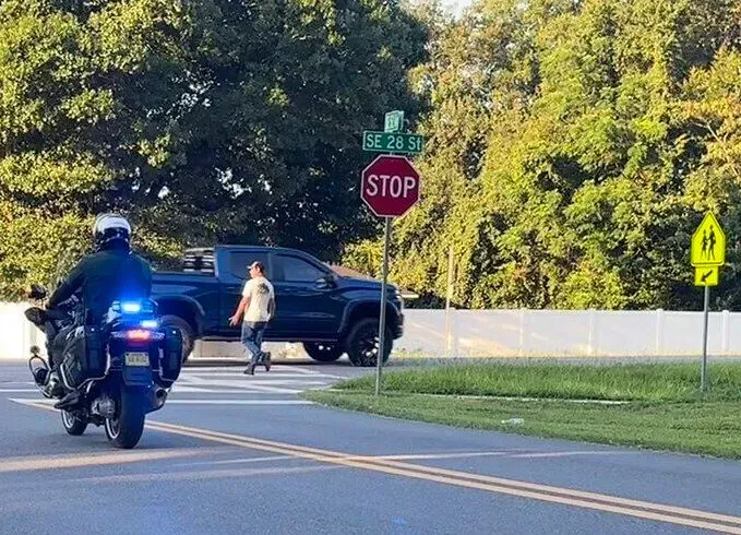 Police motorcycle and pickup truck at an intersection. A person crosses the street. Stop sign visible.