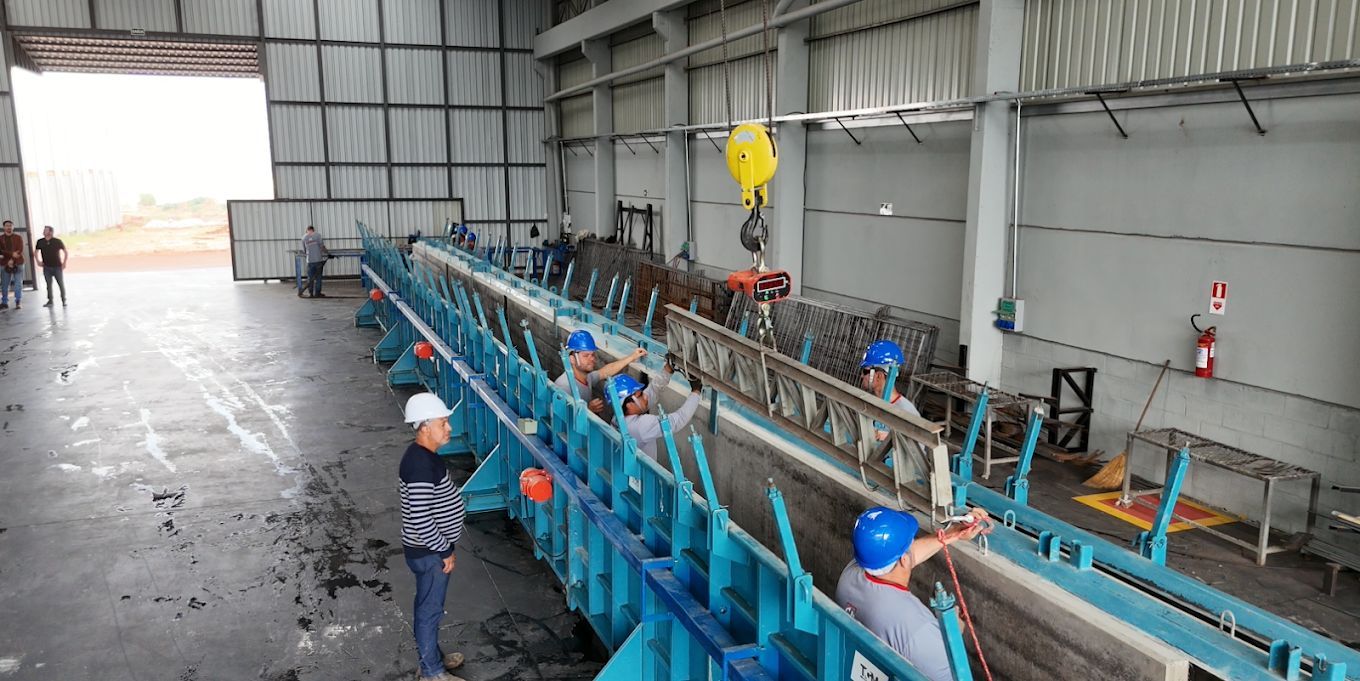 Operários com capacetes de segurança montam uma grande estrutura metálica azul dentro de um galpão bem iluminada.