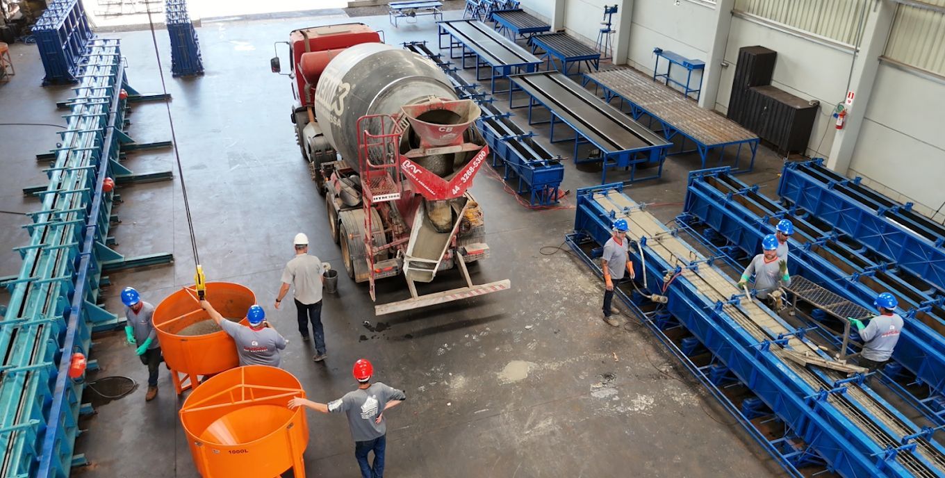Operários usam baldes laranja e pontes rolantes para transferir concreto de um caminhão betoneira para longas fôrmas azuis.