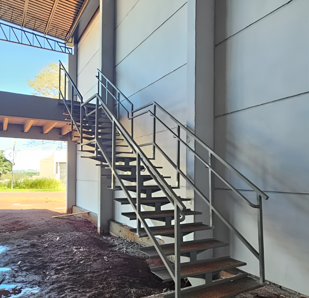 Escada metálica industrial com corrimão leva a uma plataforma elevada encostada em uma parede de concreto.