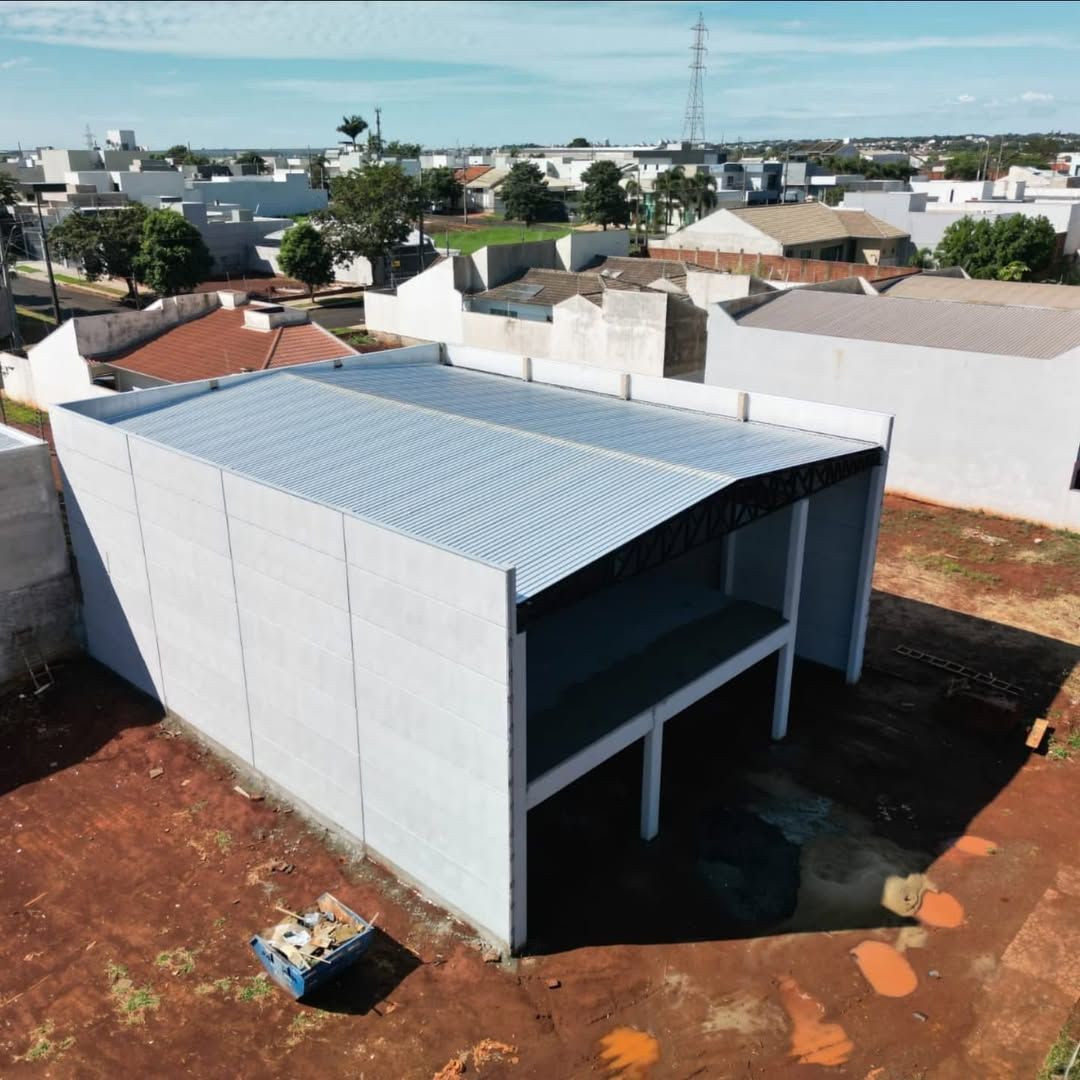 Vista aérea de uma obra retangular com paredes de concreto cinza e telhado de metal ondulado em construção.