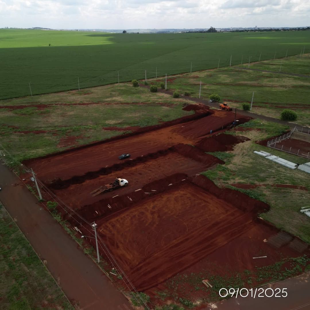 Vista aérea de um canteiro de obras com terra vermelha, máquinas pesadas e um campo rural ao fundo.