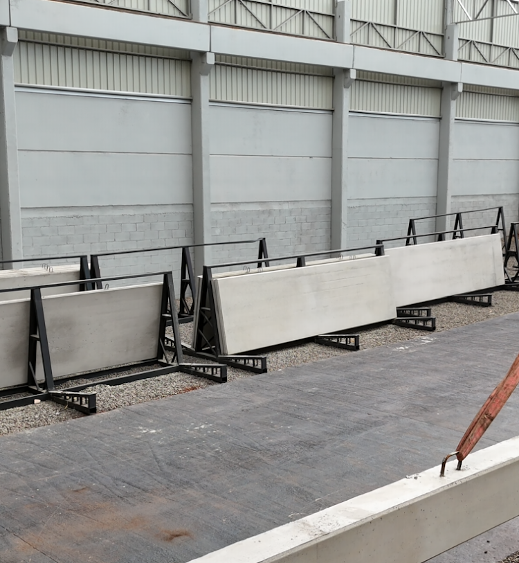 Placas retangulares de concreto armazenadas na vertical em estruturas metálicas pretas em forma de A ao lado de uma parede.