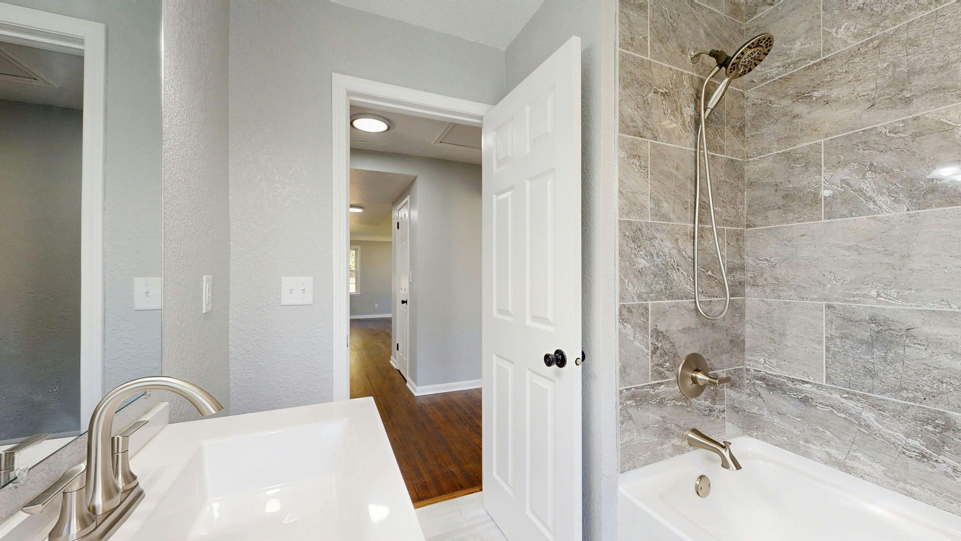 Salle de bains avec baignoire blanche, douche en marbre gris et porte ouverte sur un couloir avec parquet.