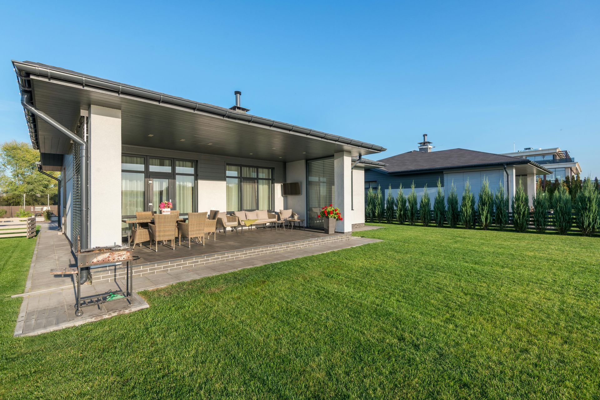 Patio de maison moderne avec sièges extérieurs donnant sur une grande pelouse verte sous un ciel bleu clair