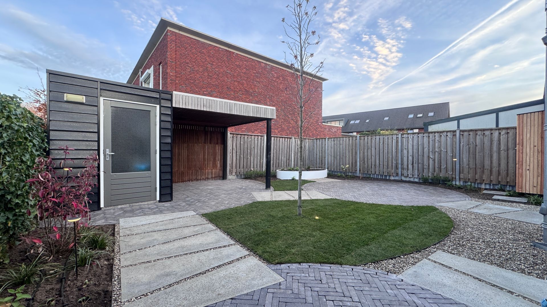 Achtertuin met schuur, houten luifel, grasveldje, bestraat pad en houten schutting tegen een bakstenen gebouw.