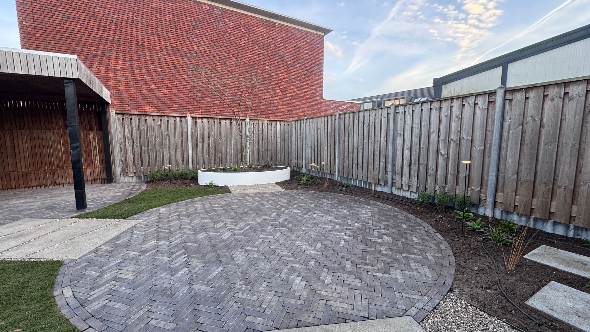 Een betegeld terras in een achtertuin met een houten schutting en een bakstenen gebouw op de achtergrond.