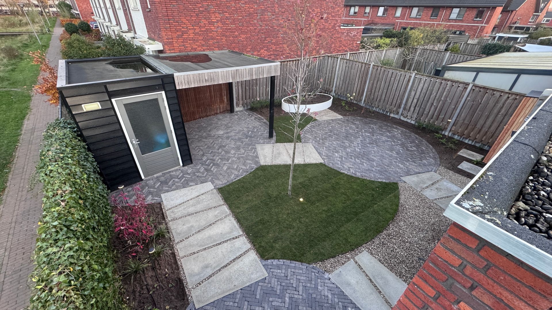 Achtertuin met betegeld terras, gazon en schuur, van bovenaf gezien. Bakstenen muur aan de rechterkant.