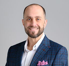 Man with beard smiling, wearing blue plaid blazer, white shirt; gray background.