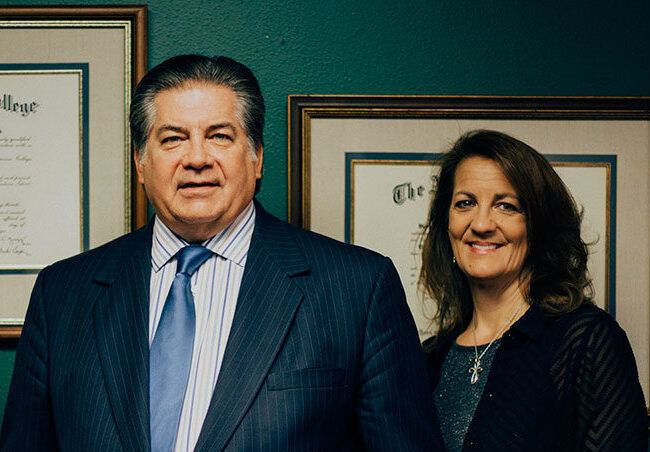 Man and woman in a law office, posing in front of framed documents. Man in suit, woman smiling.