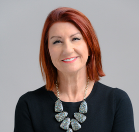 Woman with red hair wearing a black top and statement necklace smiles at the camera.