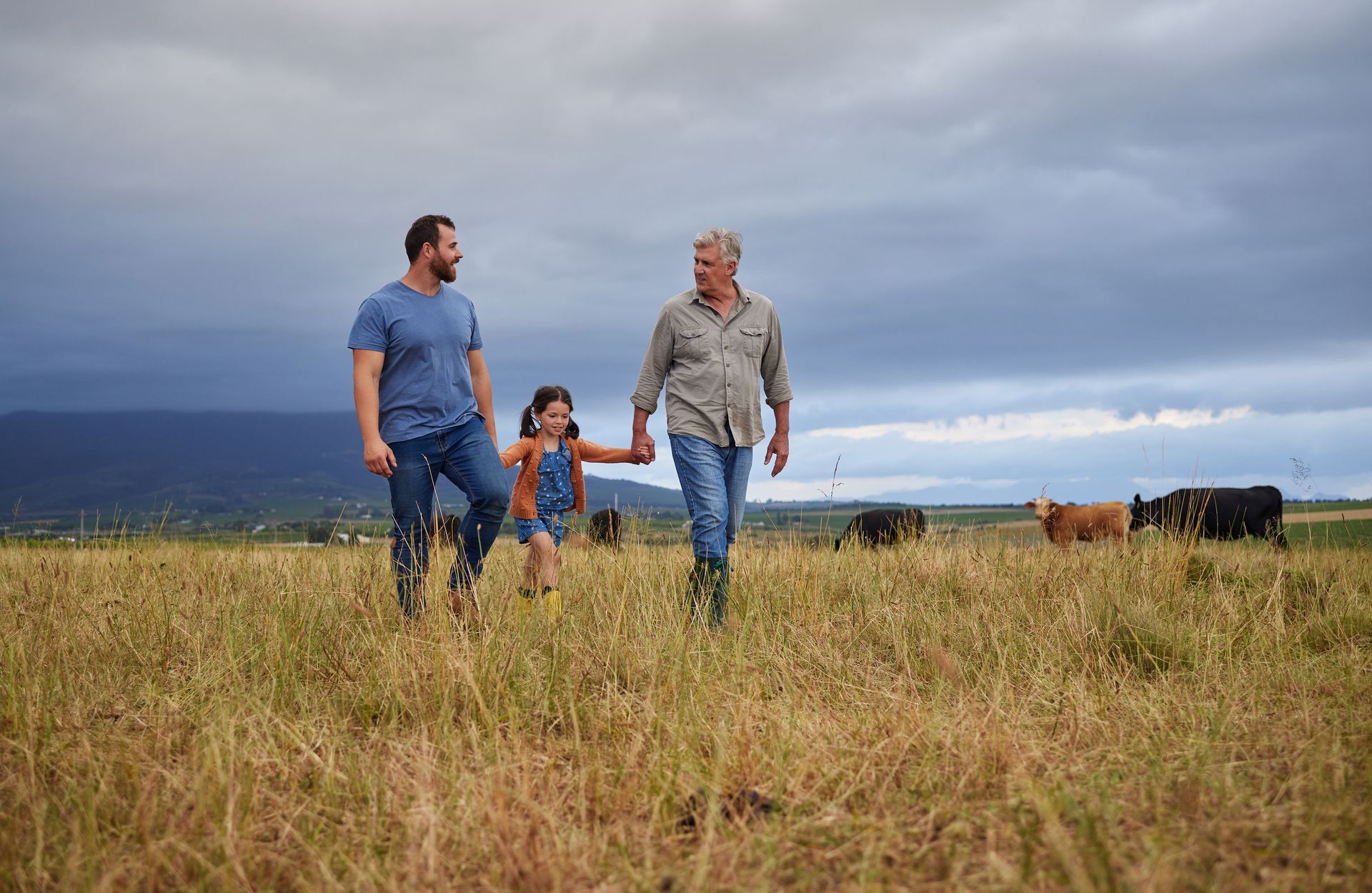 Two men and a girl walk through a field, holding hands, with cows grazing in the background under a cloudy sky.