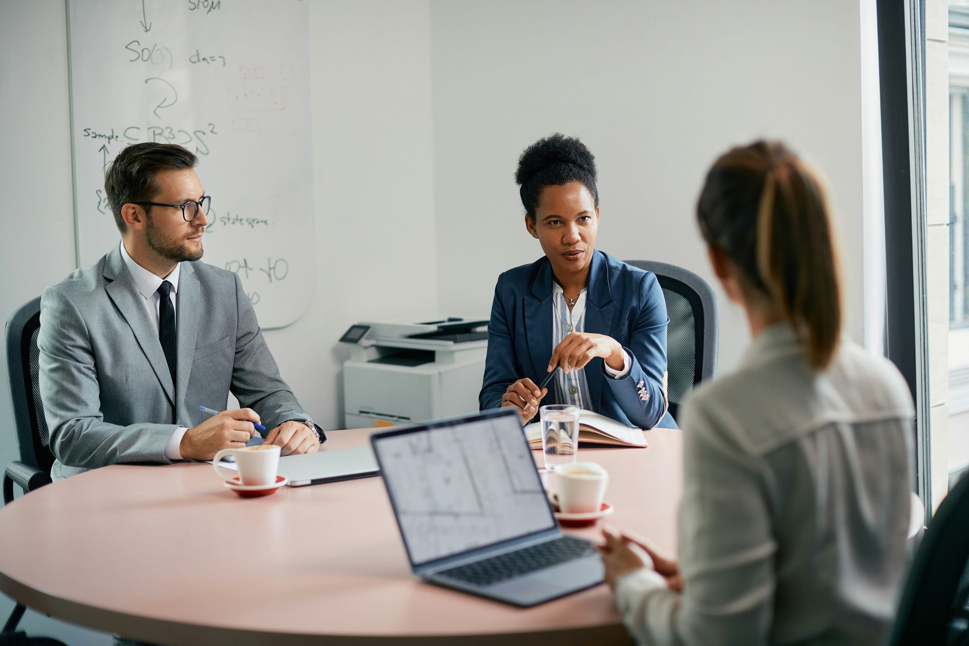 Three people in suits at a meeting in a brightly lit room; a woman is speaking, others listening.