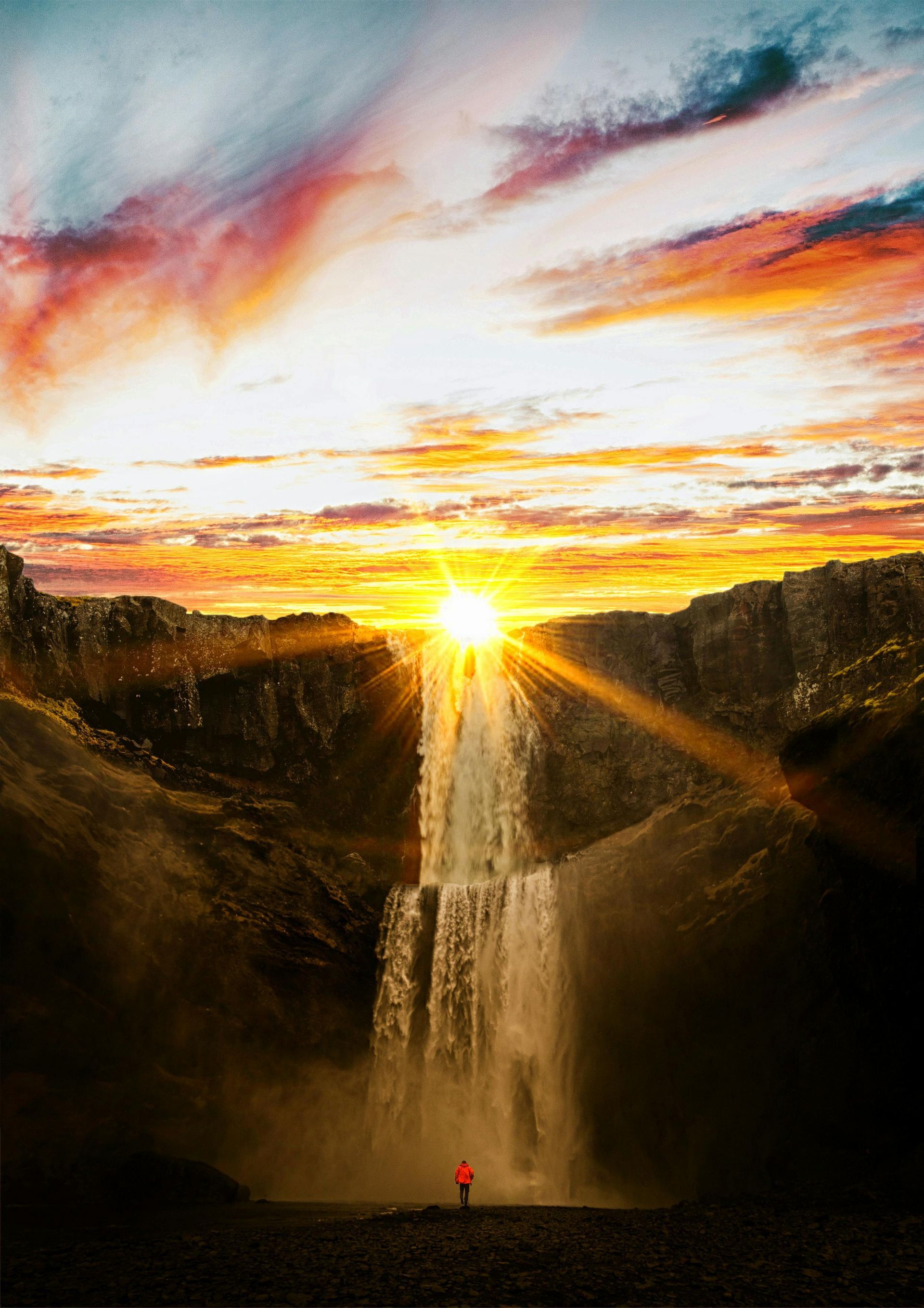 Waterfall cascading into a valley at sunset. Golden light streams from sun, illuminating a person in red.