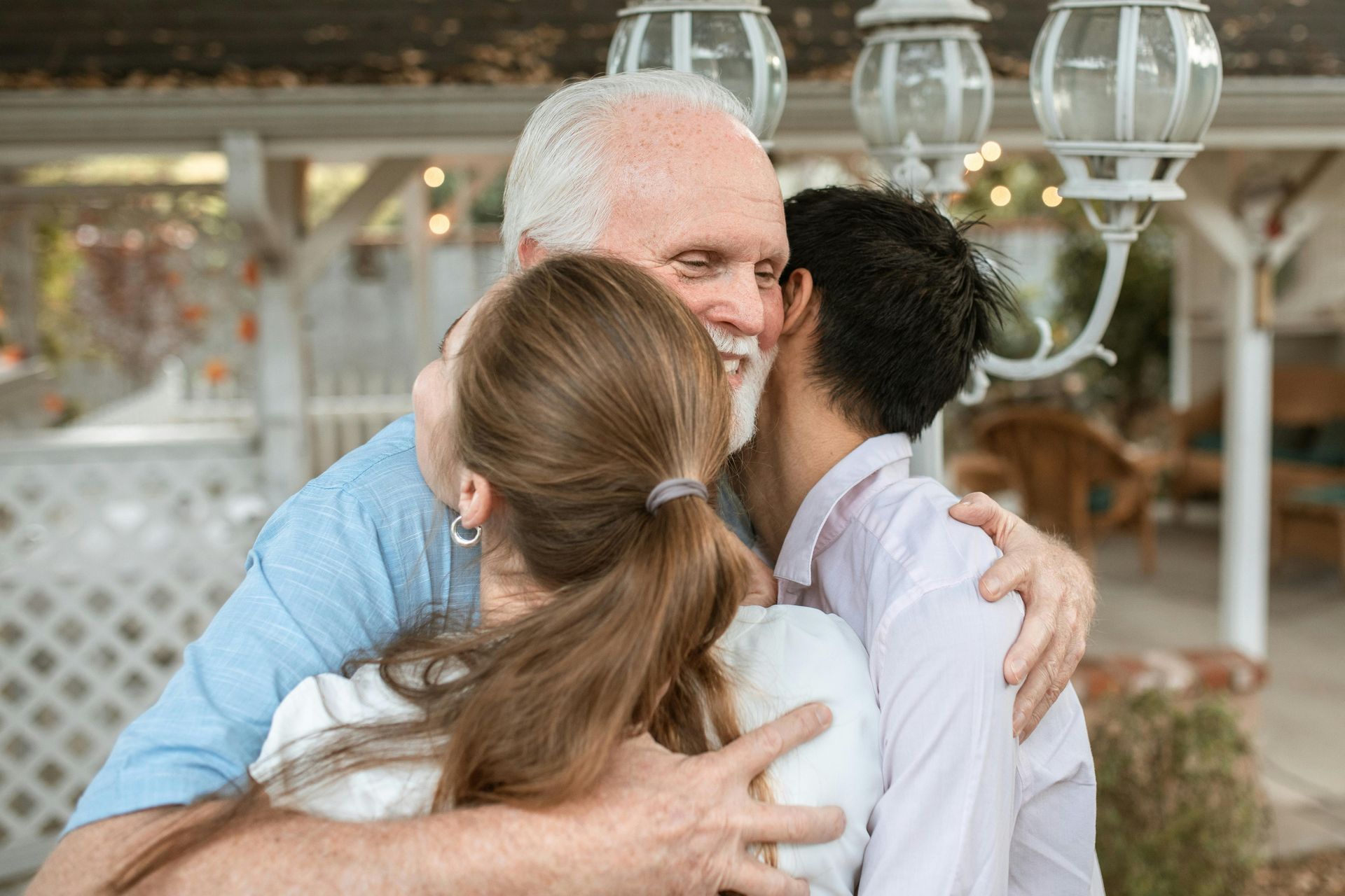 An elderly man hugs two younger people outdoors, smiling happily.