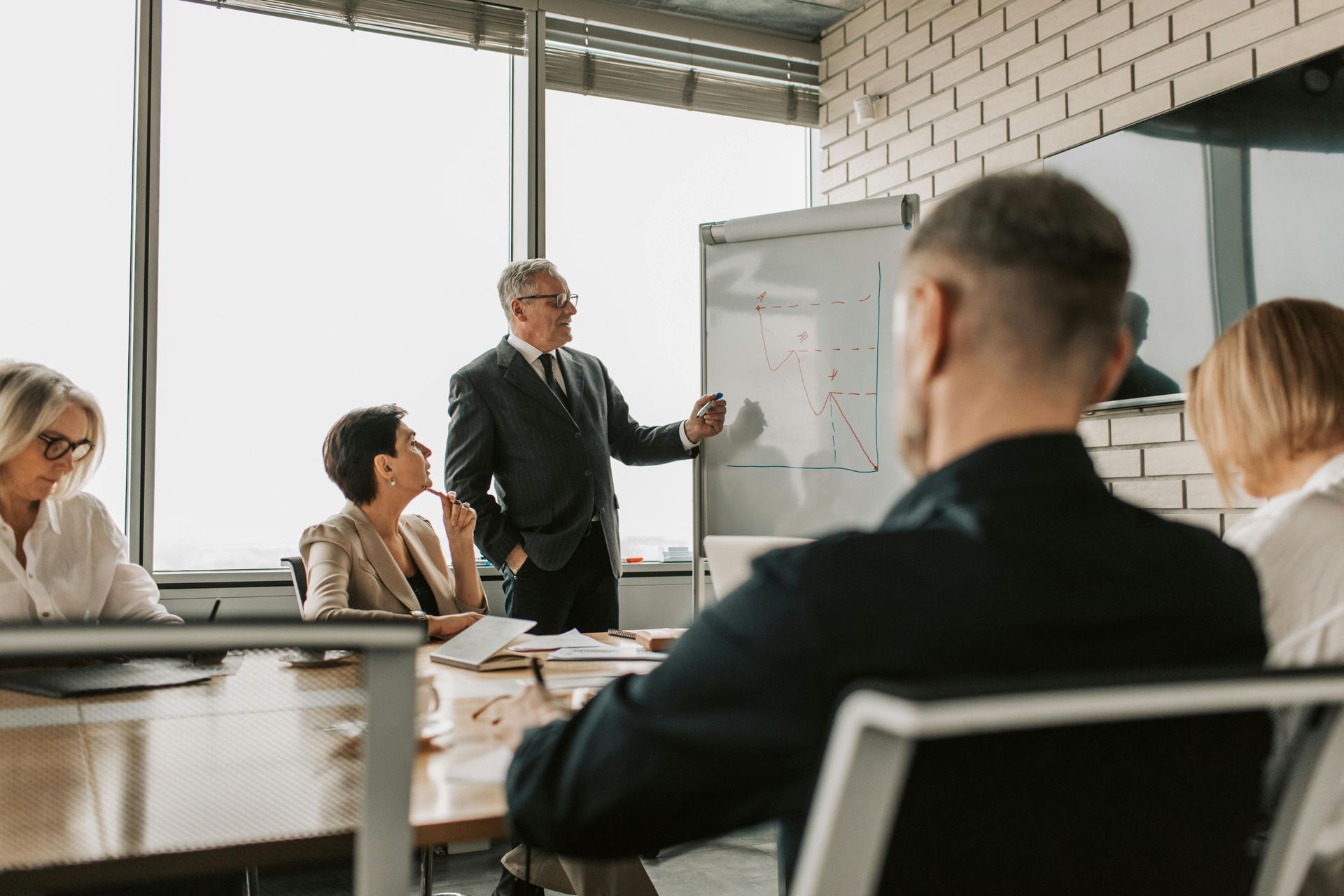 Man presenting at a meeting, pointing to a chart on a whiteboard. Several people are seated around a table.