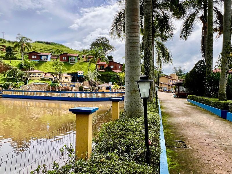 Lagoa com um caminho ao lado, ladeado por palmeiras e uma cerca baixa. Edifícios se erguem em uma colina verde ao fundo.
