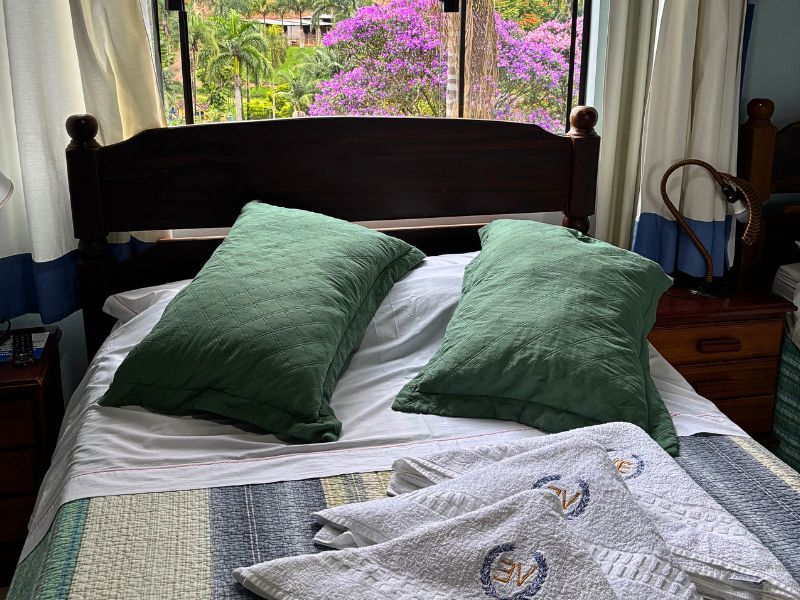 Cama com travesseiros verdes, lençóis brancos e uma janela com flores roxas ao fundo.