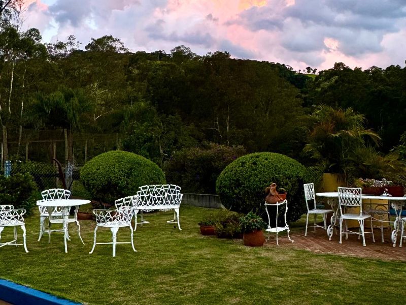 Móveis de jardim brancos em um gramado, cercado por vegetação, com uma floresta ao fundo sob um céu colorido.