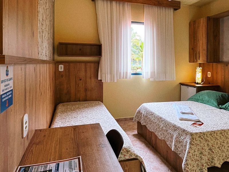 Quarto com duas camas, painéis de madeira, janela com cortinas e mesa de cabeceira com abajur.