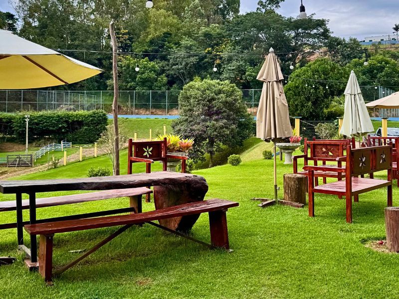 Área de estar externa com mesas de piquenique, bancos e guarda-sóis sobre grama verde, com árvores ao fundo.