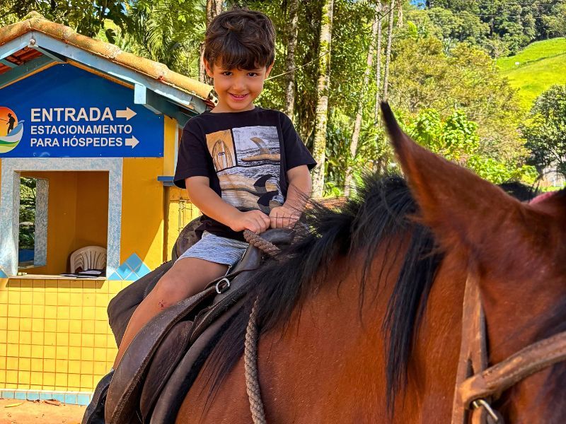Menino a cavalo sorrindo, segurando as rédeas, perto de um prédio amarelo e azul em um ambiente natural.