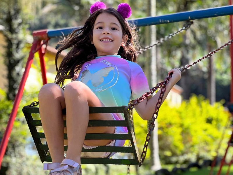 Menina sorrindo em um balanço, usando uma camisa colorida e uma tiara de pompons, ao ar livre com árvores ao fundo.