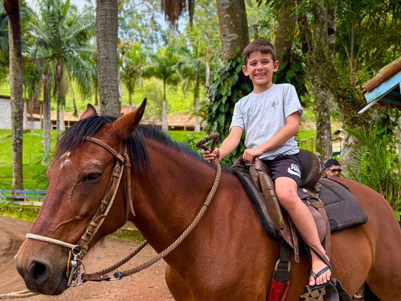 Menino sorrindo em um cavalo marrom ao ar livre, vestindo shorts e sandálias. Árvores e prédios ao fundo.