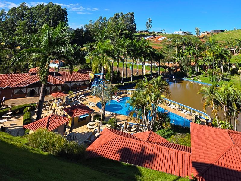 Resort com piscina, edifícios com telhados vermelhos, vegetação exuberante e um lago sob um céu azul.