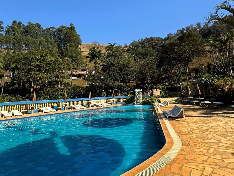 Piscina com espreguiçadeiras, árvores e colinas sob um céu azul.