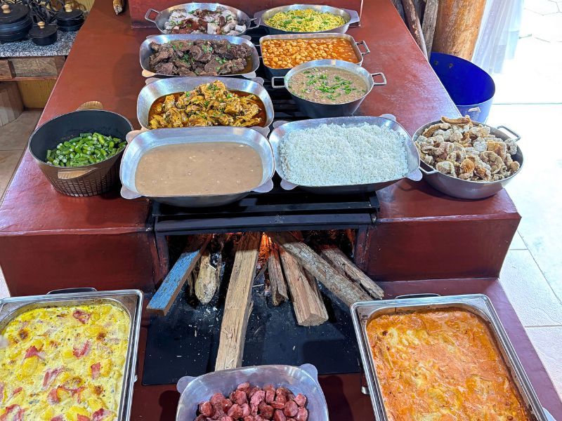 Buffet com diversos pratos preparados em um fogão a lenha com chamas. Legumes, carnes e caçarolas expostos.