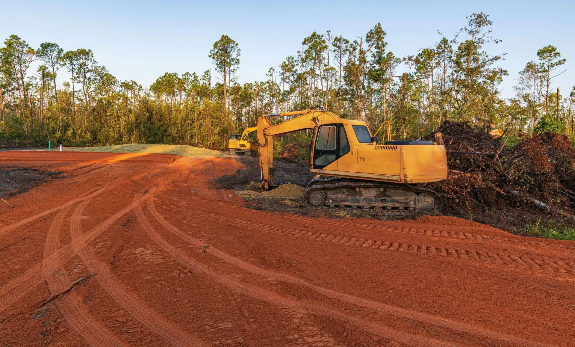Yellow excavator on a red dirt road, clearing land near a treeline.