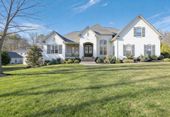 A white, multi-gabled suburban house with dark shutters and a central arched entryway sits on a large, grassy lawn.