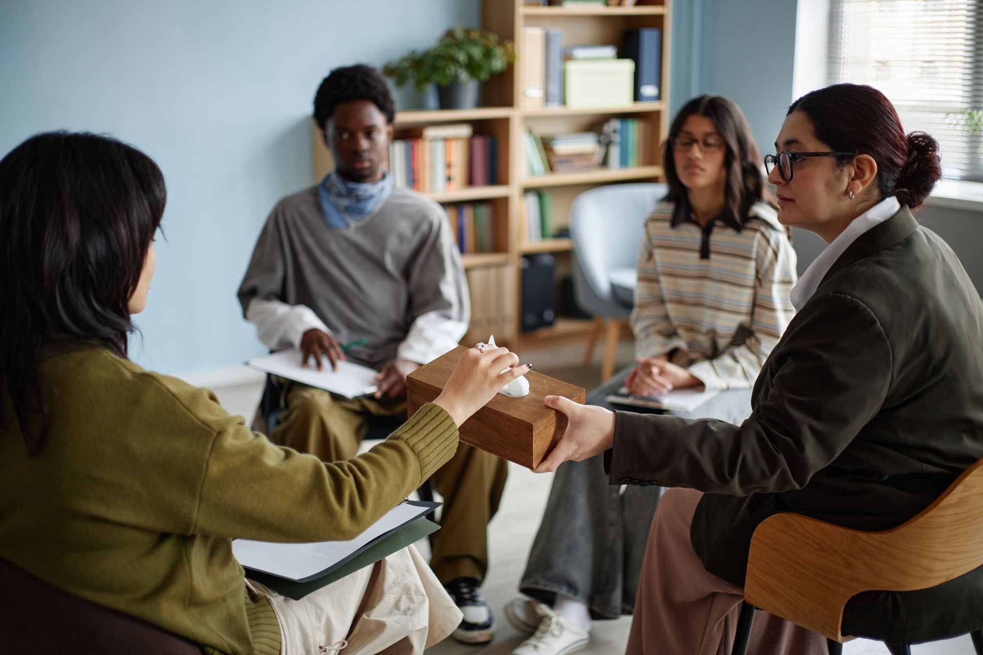 Group therapy session: diverse people seated in a circle, counselor passes tissues to a woman.