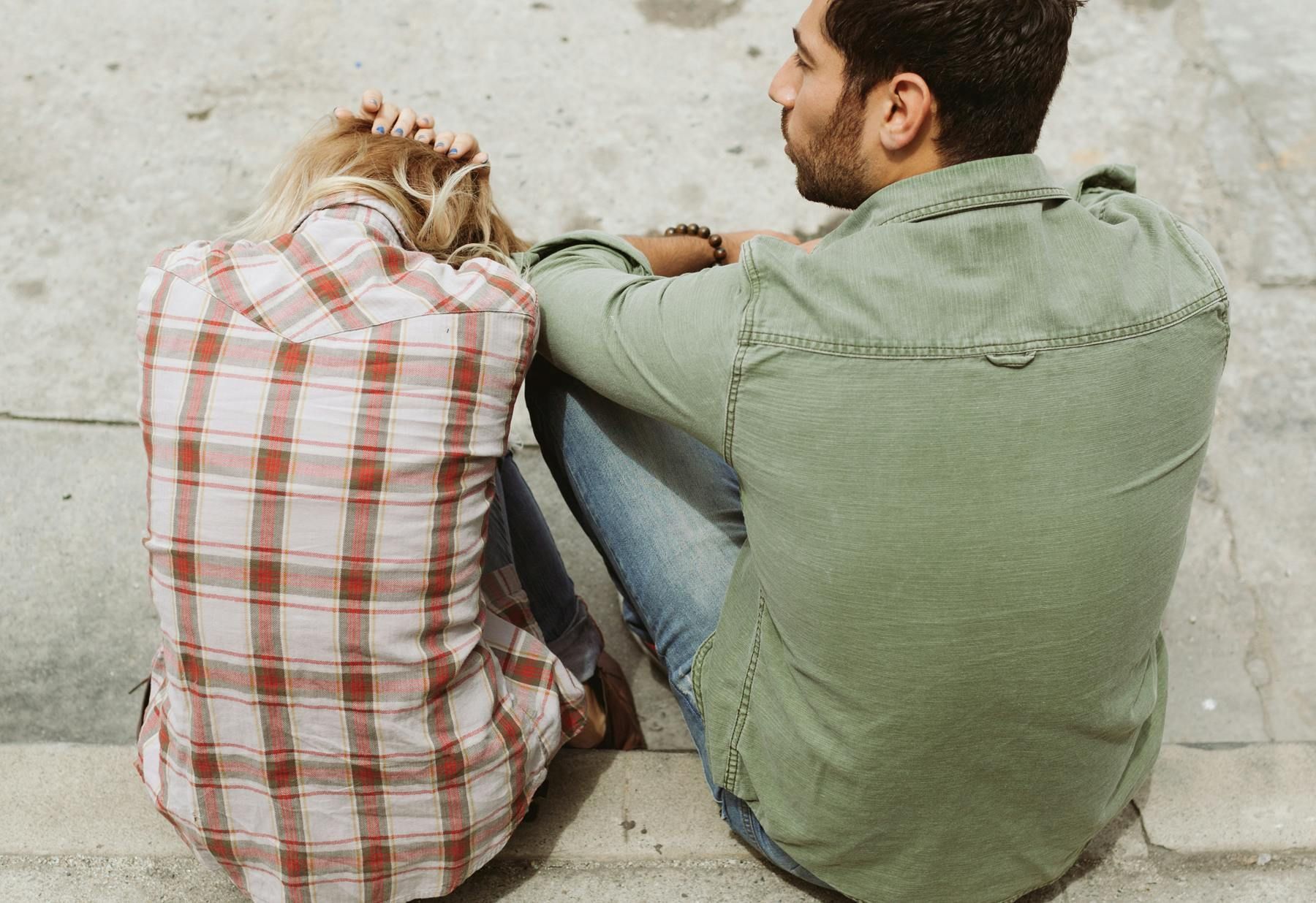 A man and a woman are sitting on the sidewalk with their backs to the camera.