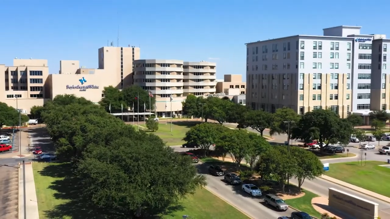 An outdoor view of a hospital complex with multiple multi-story buildings, trees, and parking areas on a sunny day.