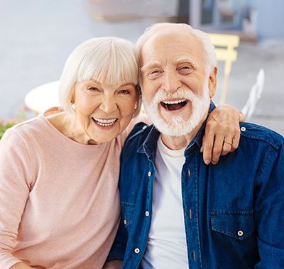 Smiling couple, woman in pink, man in denim, arm around her, outdoors.
