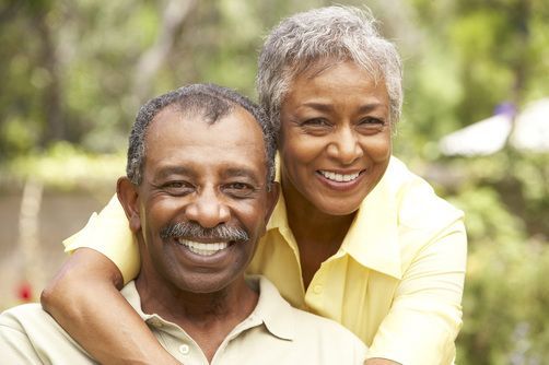 Smiling couple embracing outdoors. Woman's arms around man's shoulders, both wearing yellow shirts, green background.