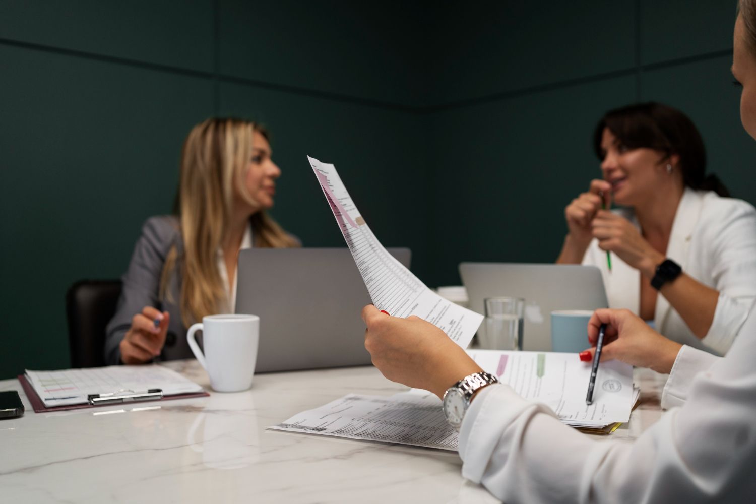 Three people in a meeting, reviewing documents. One holds papers, two look on. Laptops and mugs on a white table.