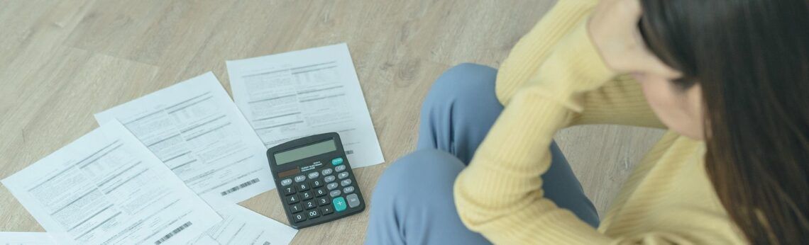 Woman with head in hands, surrounded by papers and calculator, likely stressed about finances.